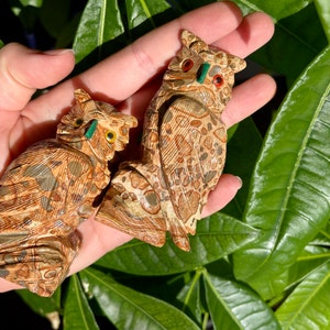 May include: Two intricately carved owl figurines made of brown and beige stone with green accents in their eyes. The owls are perched on a hand.