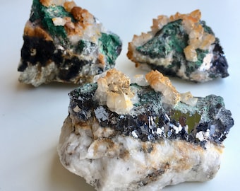 May include: Three rough, natural mineral specimens with a variety of crystals. The largest specimen has a dark gray base with a layer of shiny, metallic silver crystals. The top of the specimen is covered in small, clear quartz crystals and green malachite crystals. The other two specimens are similar in appearance, with a dark gray base and a layer of green malachite crystals.