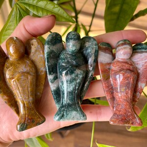 May include: Three carved angel figurines made of different types of stone. The angel on the left is brown and gold, the middle angel is green and white, and the angel on the right is red and white.