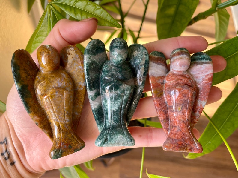 May include: Three carved angel figurines made of different types of stone. The angel on the left is brown and gold, the middle angel is green and white, and the angel on the right is red and white.