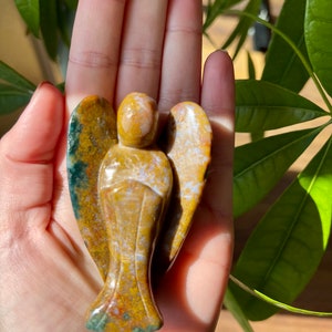 May include: A carved angel figurine made of brown, yellow, and green jasper. The angel has wings and a long flowing robe.