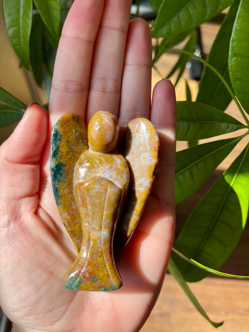 May include: A carved angel figurine made of brown, yellow, and green jasper. The angel has wings and a long flowing robe.