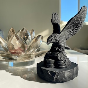 May include: A black eagle sculpture perched on a stack of coins, with its wings spread wide. The eagle is perched on a black rock base.