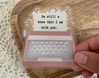 Puede incluir: Una máquina de escribir rosa con un diseño floral y el texto "Be still & know that I am with you."