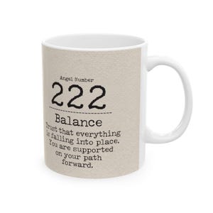 May include: White ceramic mug with a beige textured design. The mug features the text "Angel Number 222 Balance" in black, along with an inspirational message. The mug has a white handle and is set against a white background.
