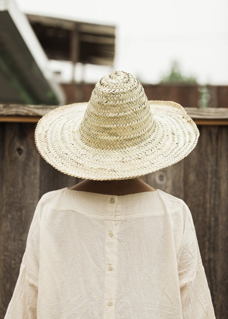 May include: A person wearing a large, wide-brimmed straw hat with a pointed crown. The hat is made of woven straw and has a natural color. The person is wearing a white, button-down shirt.