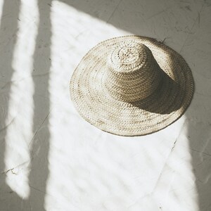 May include: A wide-brimmed straw hat with a woven texture sits on a white surface. The hat is in focus, while the background is blurred.