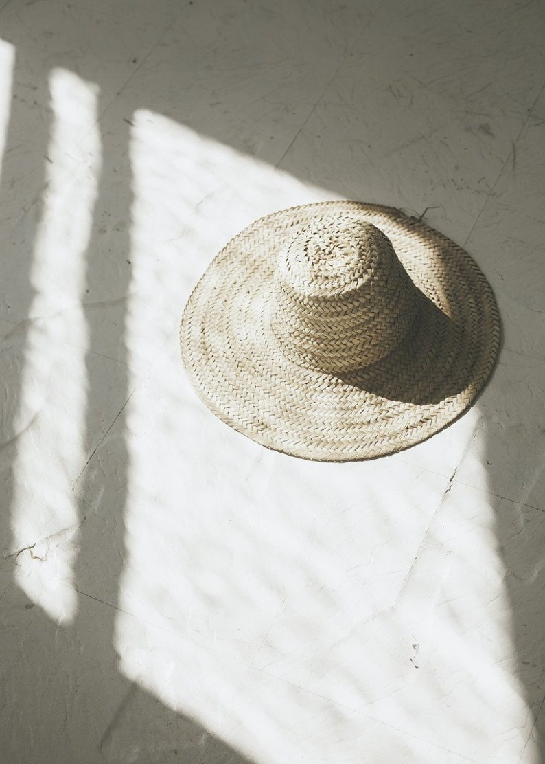 May include: A wide-brimmed straw hat with a woven texture sits on a white surface. The hat is in focus, while the background is blurred.