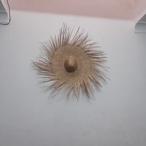 K&ouml;nnte beinhalten: Ein gro&szlig;er, runder, geflochtener Strohhut mit breiter Krempe, der an einer wei&szlig;en Wand h&auml;ngt.