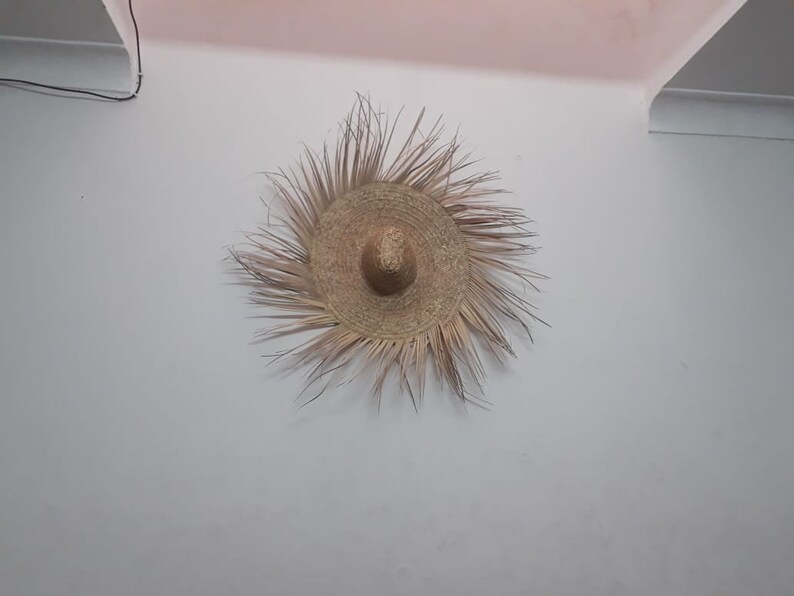K&ouml;nnte beinhalten: Ein gro&szlig;er, runder, geflochtener Strohhut mit breiter Krempe, der an einer wei&szlig;en Wand h&auml;ngt.