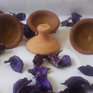 Puede incluir: Tres pequeñas macetas de terracota con base redonda y una abertura ancha y poco profunda. Las macetas están dispuestas sobre una superficie blanca con pétalos de flores moradas esparcidos a su alrededor.