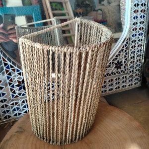 May include: A woven lampshade made of natural fibers, with a cylindrical shape and a light brown color. The lampshade is made of a series of vertical strands of rope, creating a textured and organic look.
