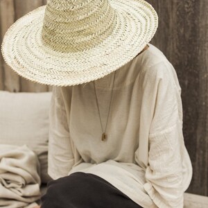 May include: A person wearing a large, wide-brimmed straw hat with a high crown. The hat is made of woven straw and has a natural color. The person is wearing a white linen shirt and black pants. They are sitting on a wooden surface.