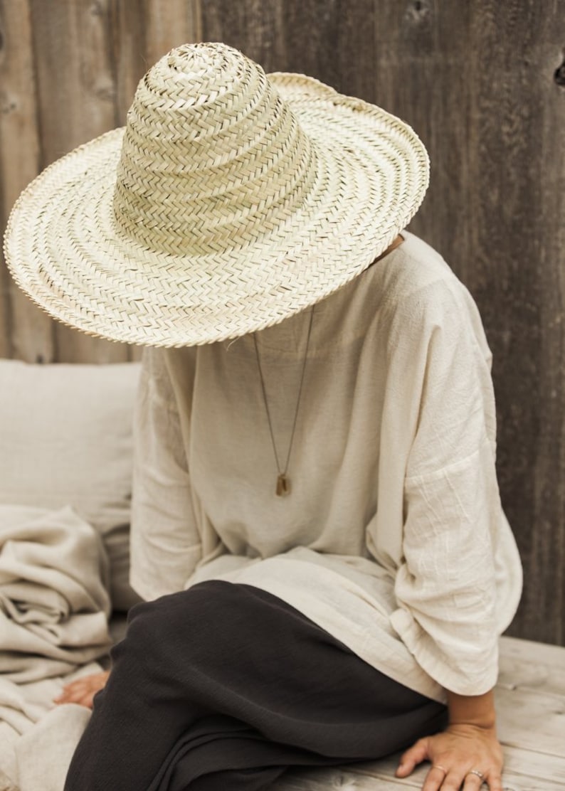 May include: A person wearing a large, wide-brimmed straw hat with a high crown. The hat is made of woven straw and has a natural color. The person is wearing a white linen shirt and black pants. They are sitting on a wooden surface.