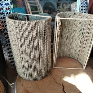 May include: Two cylindrical woven lampshades made of natural fiber rope. The lampshades are beige and have a natural, rustic look.