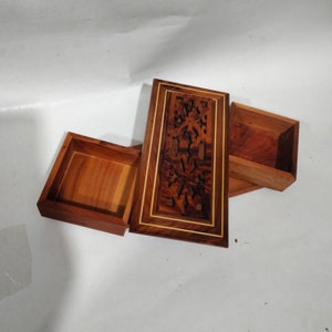 Op de afbeelding: Een houten doos met ingewikkelde houtsnijwerk en een schuifdeksel. De doos is gemaakt van donker hout met lichte hout accenten. De doos is open en het deksel is naar de zijkant geschoven.