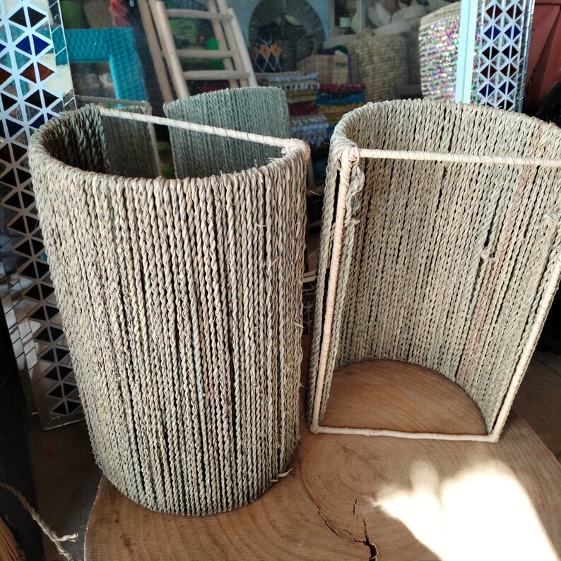 May include: Two woven lampshades made of natural materials, one cylindrical and one rectangular, sitting on a wooden surface.