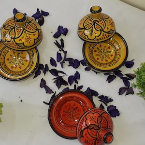 May include: Two sets of Moroccan ceramic serving dishes with lids. The dishes are yellow with red and black floral designs. The lids are dome-shaped and have a knob on top. The dishes are on a white surface with purple flower petals.