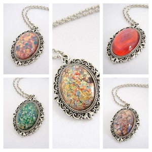 May include: Four silver necklaces with oval-shaped pendants featuring different colored glass stones. The stones have a marbled or speckled appearance. The necklaces are on a white background.