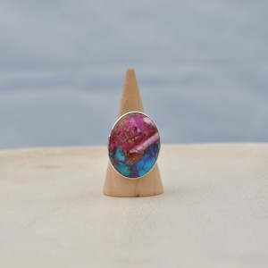 Könnte beinhalten: Ein silberner Ring mit einem großen ovalen Stein. Der Stein ist eine Mischung aus rosa, blau und goldenen Farben.
