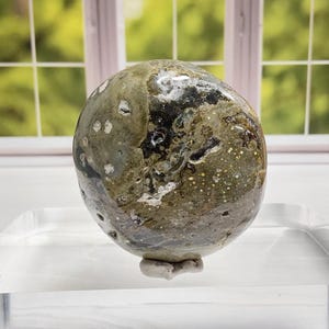 May include: A polished, rounded stone with a mottled pattern of green, gray, and black. The stone has a smooth, glossy surface and sits on a small, light-colored base. The background features a window with a view of green foliage.