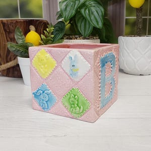 May include: A square pink ceramic planter featuring a quilted texture with embossed diamond-shaped panels. Each panel displays a raised pastel animal figure in yellow, white, blue, or green. A large blue letter B is embossed on the side.