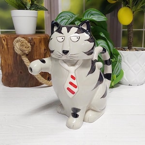 May include: A ceramic teapot shaped like a sitting cat with gray and black stripes. The cat features a white tie with red diagonal stripes, pointed ears, and a spout extending from its right side. The handle is formed by the cat's curved tail.