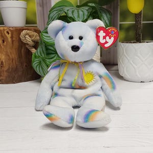 May include: A plush bear toy featuring a white base with pastel rainbow tie-dye patterns. The bear has a yellow ribbon tied around its neck, a yellow embroidered sun on its chest, and a red heart-shaped Ty Beanie Baby tag attached to its ear.