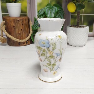 May include: A white porcelain vase with a scalloped rim and gold trim, featuring a delicate botanical print of blue, yellow, and white wildflowers with green leaves and small insects scattered across the surface on a white background.