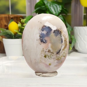 May include: A smooth, oval-shaped polished stone with a pale pink and cream base color. The surface features a prominent, irregular dark blue and black dendritic inclusion in the center, surrounded by subtle patches of earthy green and white mineral deposits.