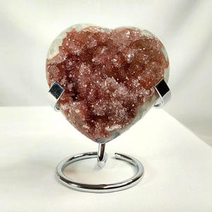 May include: A heart-shaped amethyst geode with a deep reddish-purple crystal interior. The geode is mounted on a silver-colored metal stand. The stand has a circular base and two curved supports that cradle the geode.
