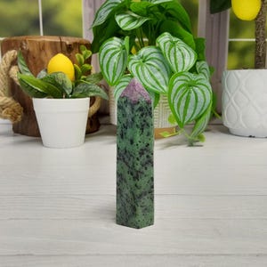 May include: A tall, rectangular gemstone tower. The stone is a deep green with black speckles and a pinkish-purple tip. The polished crystal is standing upright, showcasing its natural patterns and colors. The background includes plants and a wooden element.