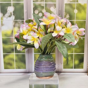 May include: A bouquet of artificial plumeria flowers in a decorative vase. The flowers have yellow centers, white and pink petals, and green leaves. The vase is a ribbed design with shades of green and purple.