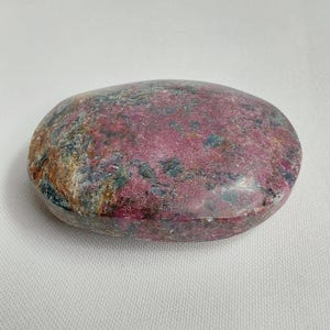 May include: A polished, oval-shaped gemstone with a mix of colors. The stone features a dominant deep pink hue, interspersed with patches of darker red, green, and brown. The surface is smooth and reflective, suggesting a high polish.