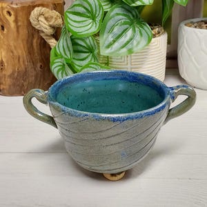 May include: A ceramic bowl with two handles and a footed base. The exterior features a light gray glaze with subtle diagonal carved lines, while the interior is finished in a vibrant teal blue glaze with a darker blue rim and speckled texture.