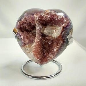 May include: A heart-shaped amethyst geode with a polished exterior and a crystalline interior. The geode is mounted on a silver-colored metal stand. The amethyst crystals display shades of purple and red.