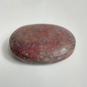 May include: A smooth, oval-shaped stone with a mottled appearance. The stone's color is primarily a deep reddish-brown, with subtle variations and hints of other colors. The surface appears polished and slightly reflective, suggesting a natural gemstone or decorative item.