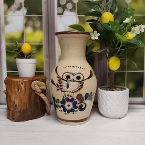 May include: A beige ceramic vase with a speckled finish, featuring a hand-painted brown owl illustration and blue and green floral motifs. The vase has a flared rim and a tapered base, with brown painted bands at the top and bottom edges.
