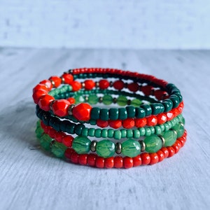 May include: A set of five beaded bracelets in red and green. The bracelets are stacked on top of each other, creating a layered look.