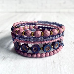 May include: A set of five beaded bracelets in shades of purple and pink. The bracelets are made with different sizes and shapes of beads, including small seed beads, larger round beads, and flower-shaped beads.
