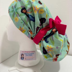May include: A light mint green scrub cap featuring a repeating pattern of cartoon fairies and yellow flowers, accented with a bright pink fabric bow at the back. The cap is displayed on a white mannequin head with a small rectangular label attached.