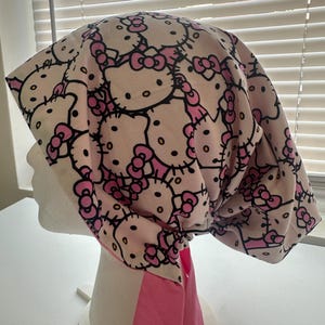 May include: A pink and white patterned surgical scrub cap featuring a repeating design of Hello Kitty characters with pink bows. The cap has a pink tie in the back.