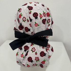 May include: A white scrub cap featuring a pattern of red ladybugs and flowers. The cap is tied with a large black bow. The ladybugs have black spots and the flowers are red with green leaves.