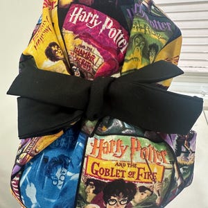 May include: A fabric scrub cap featuring a colorful collage of Harry Potter book covers, including The Chamber of Secrets and The Goblet of Fire. The cap is accented with a large, solid black fabric bow tied at the center front of the head covering.