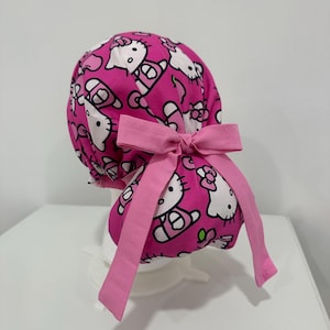 May include: A bright pink scrub cap featuring a repeating pattern of the Hello Kitty character with white faces and pink bows. The cap has a gathered back design and is secured with a solid light pink fabric ribbon tied into a bow at the center.