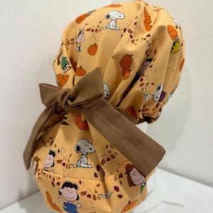 May include: A scrub cap in a light orange fabric featuring a repeating pattern of Snoopy, Charlie Brown, and other Peanuts characters amidst autumn leaves and pumpkins. The cap includes a solid brown fabric tie at the back for adjustment.