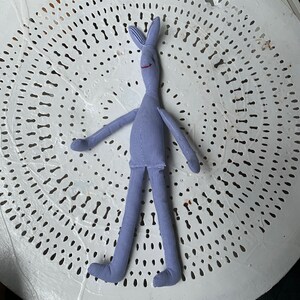 Puede incluir: Un juguete de peluche azul claro con forma de persona con brazos y piernas largos. El juguete tiene una cara sonriente con labios rojos y una nariz larga y puntiaguda.