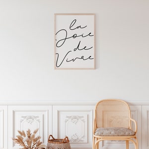 Puede incluir: Una impresi&oacute;n enmarcada con el texto "la joie de vivre" en escritura cursiva negra sobre fondo blanco. La impresi&oacute;n est&aacute; colgada en una pared blanca con z&oacute;calos blancos y una silla de mimbre con un coj&iacute;n gris claro.