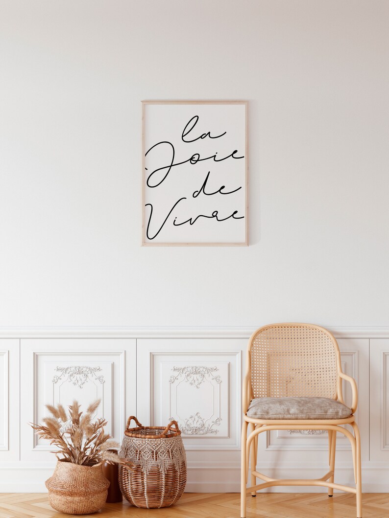 Puede incluir: Una impresi&oacute;n enmarcada con el texto "la joie de vivre" en escritura cursiva negra sobre fondo blanco. La impresi&oacute;n est&aacute; colgada en una pared blanca con z&oacute;calos blancos y una silla de mimbre con un coj&iacute;n gris claro.