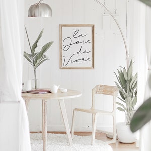Puede incluir: Una mesa redonda blanca con una silla blanca y una alfombra blanca. Un estampado enmarcado con el texto "la joie de vivre" cuelga de la pared. Hay una planta en maceta en el suelo y una l&aacute;mpara con una pantalla plateada.
